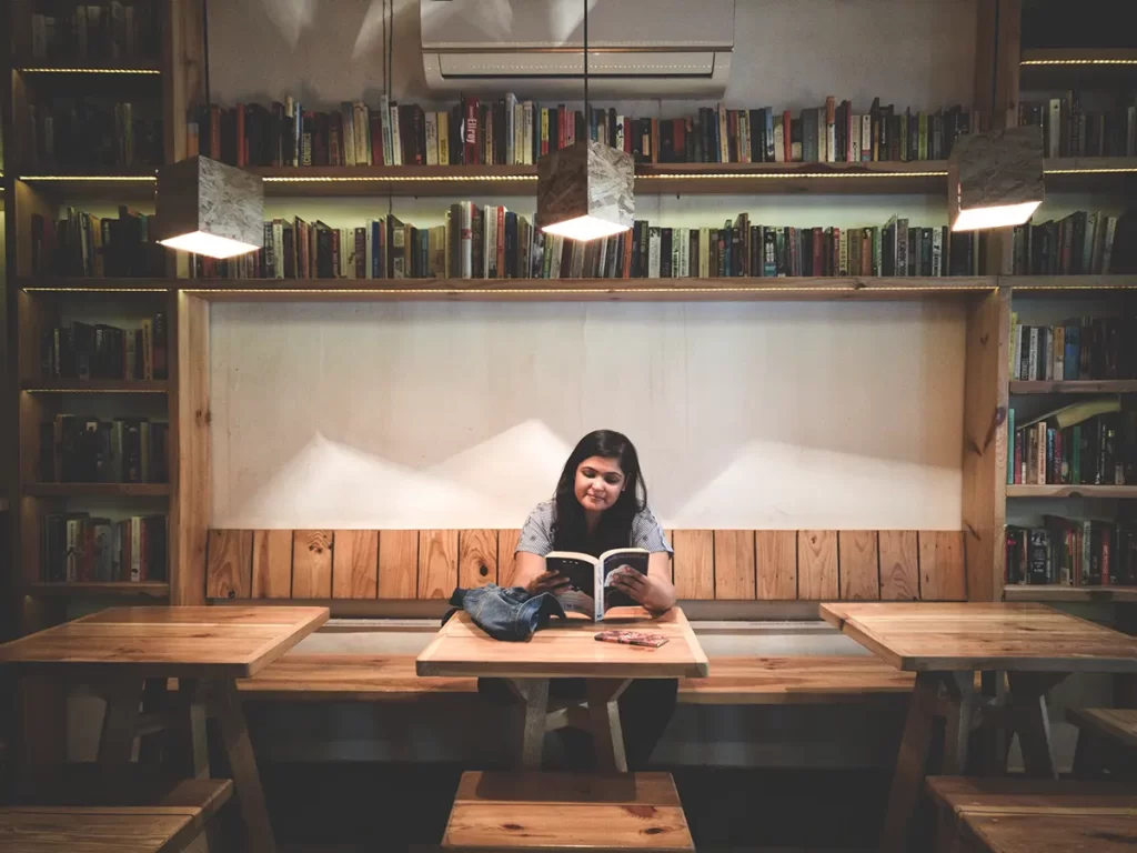 a full bookcase and a young woman sitting on a bench reading a book